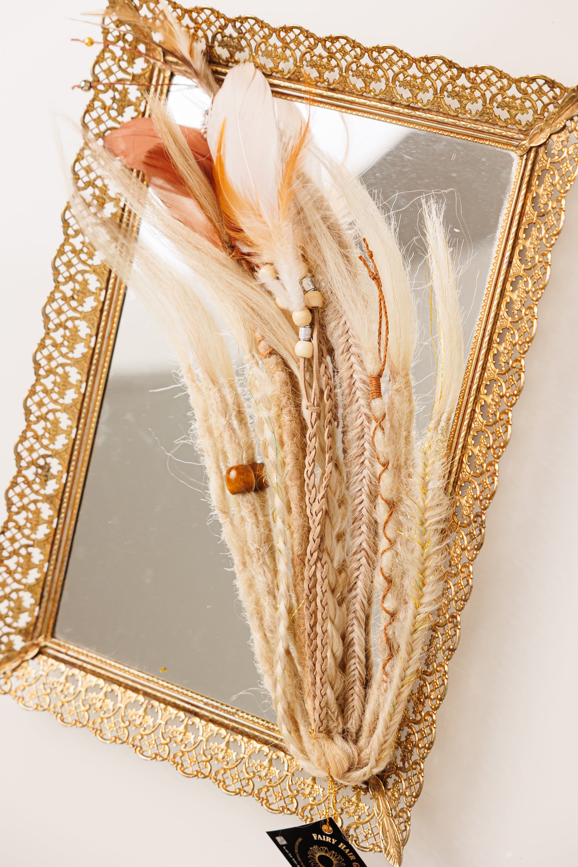 Decorative hair extension with feathers and beads in front of a gold-framed mirror.