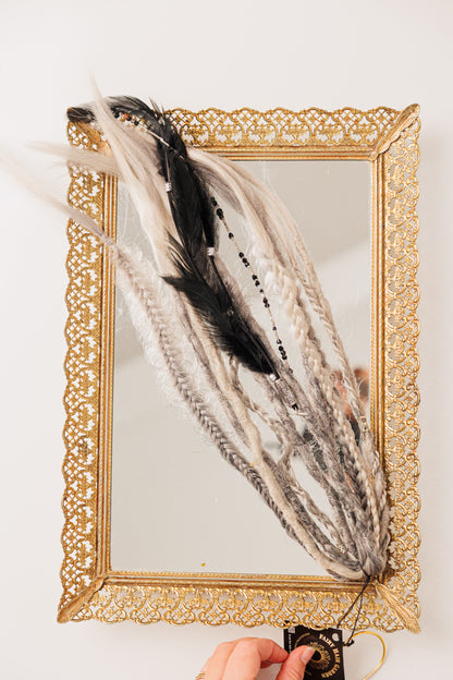 Ornate gold mirror with braided black and gray fibers, feather accent, and decorative tag.