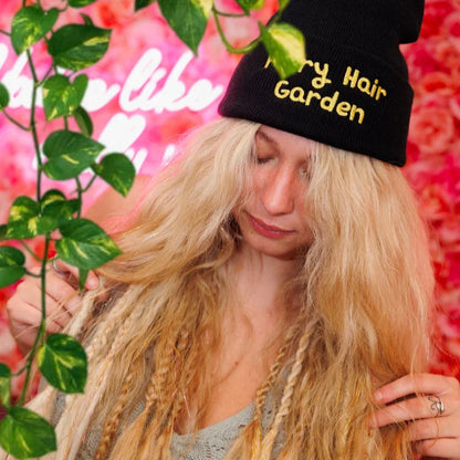 Person with wearing long boho blonde hair wearing a black beanie with 'Fairy Hair Garden' text, surrounded by green leaves and pink flowers.