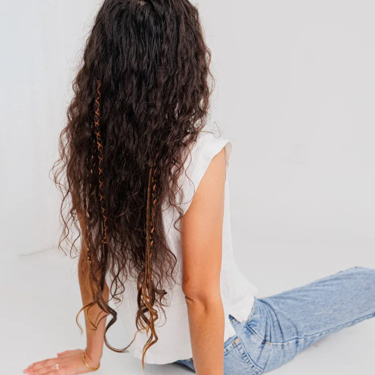 Person with long, wavy with boho hair sitting on a white floor against a white wall.