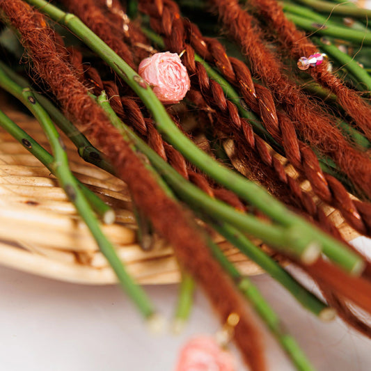 Decorative arrangement of dark red dreadlocks and green sticks with pink flower on a woven surface.