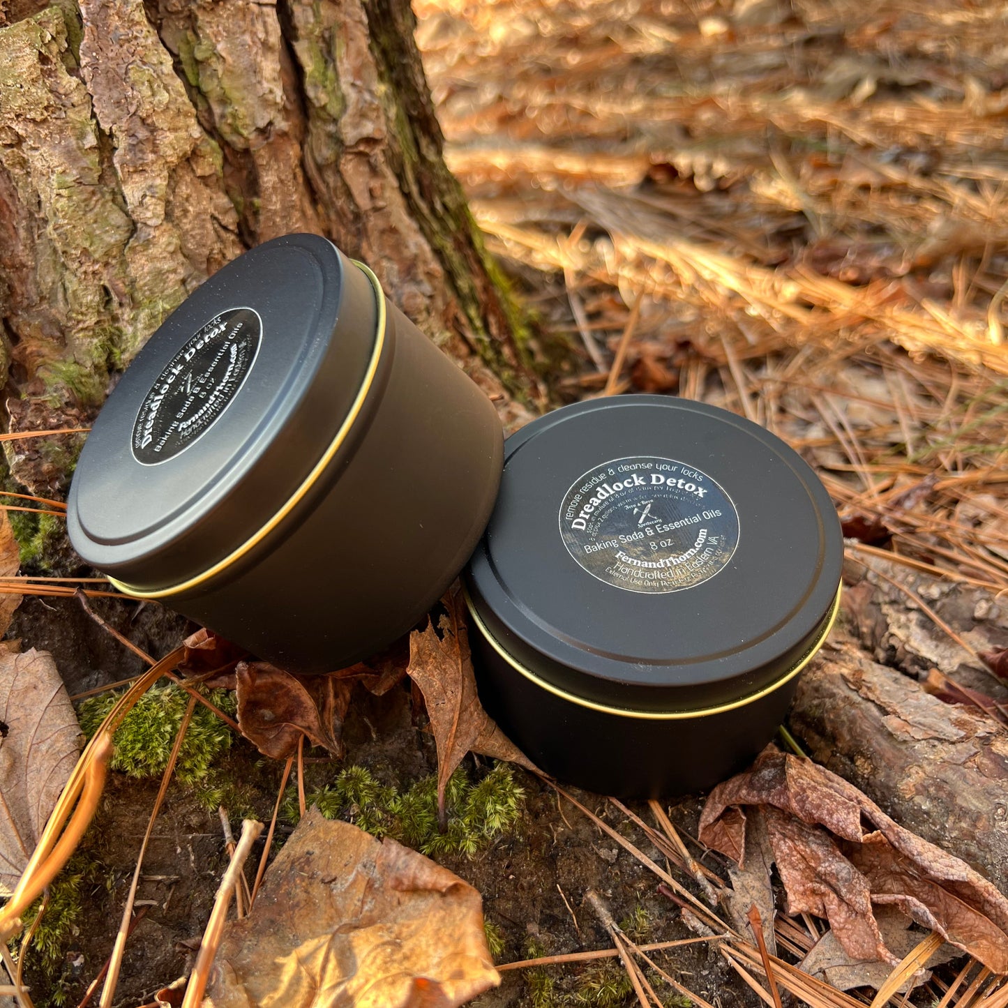 Two dreadlocks detox containers with gold accents on a forest floor.