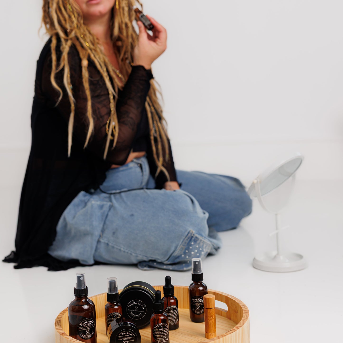 Woman with dreadlocks sitting on the floor with a tray of bottles and a white mirror in the background