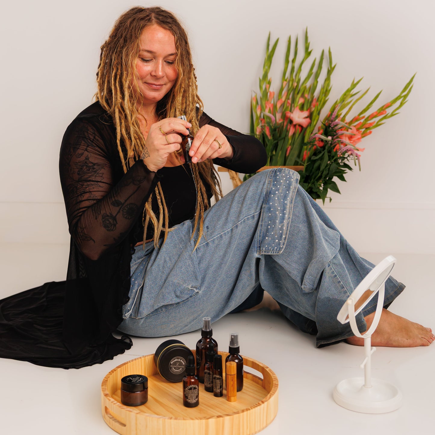 Person with dreadlocks sitting on the floor with a tray of bottles and a plant in the background