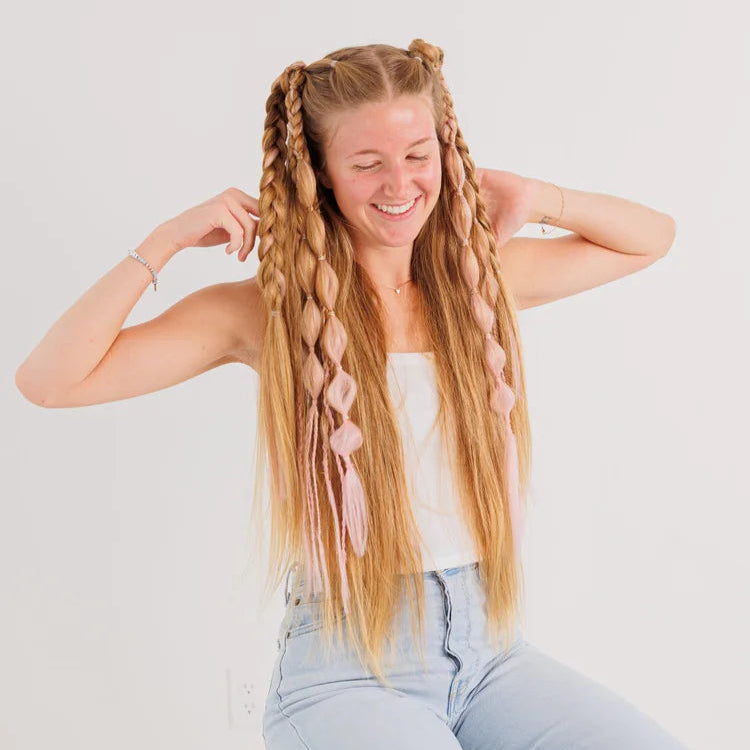 Woman with long blonde hair styled in braids, wearing a white top and light blue jeans, on a plain background.