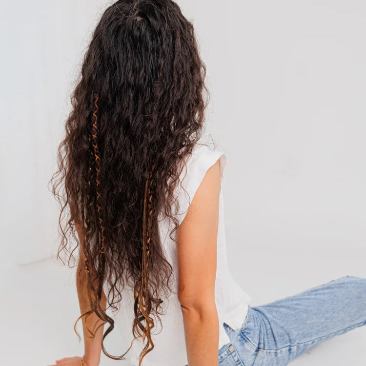 Person with long, wavy boho hair extension sitting on a white floor against a white wall.