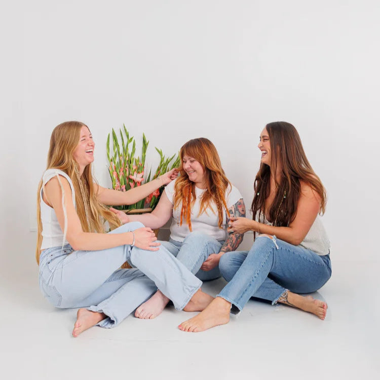 Three women wearing boho hair extension sitting on a white floor with a plant in the background