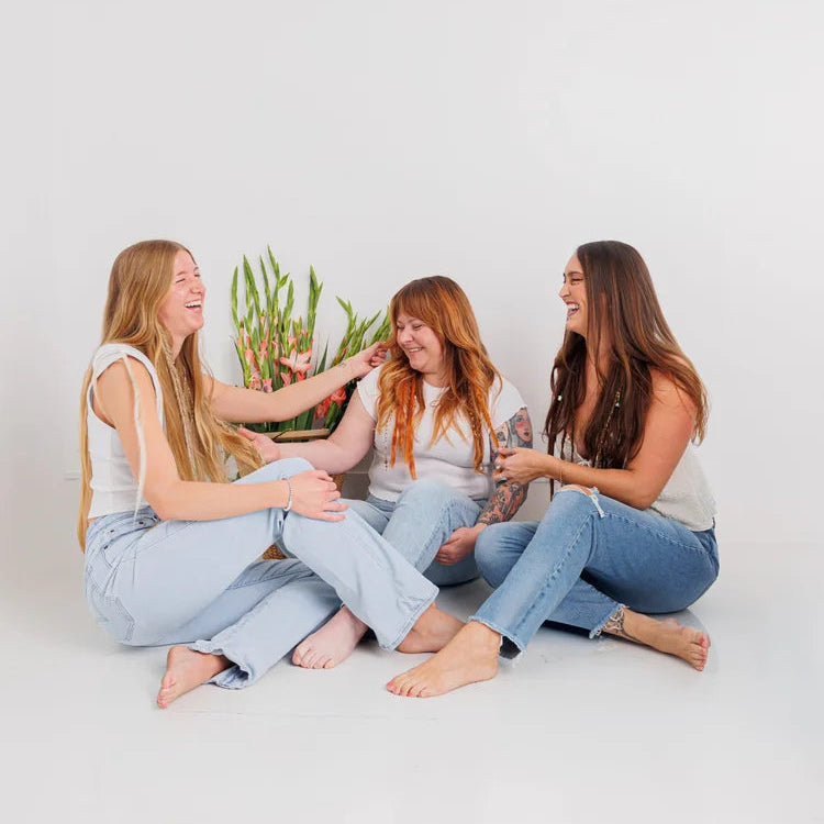 Three women sitting on a white floor with a minimal background wearing customize boho hair