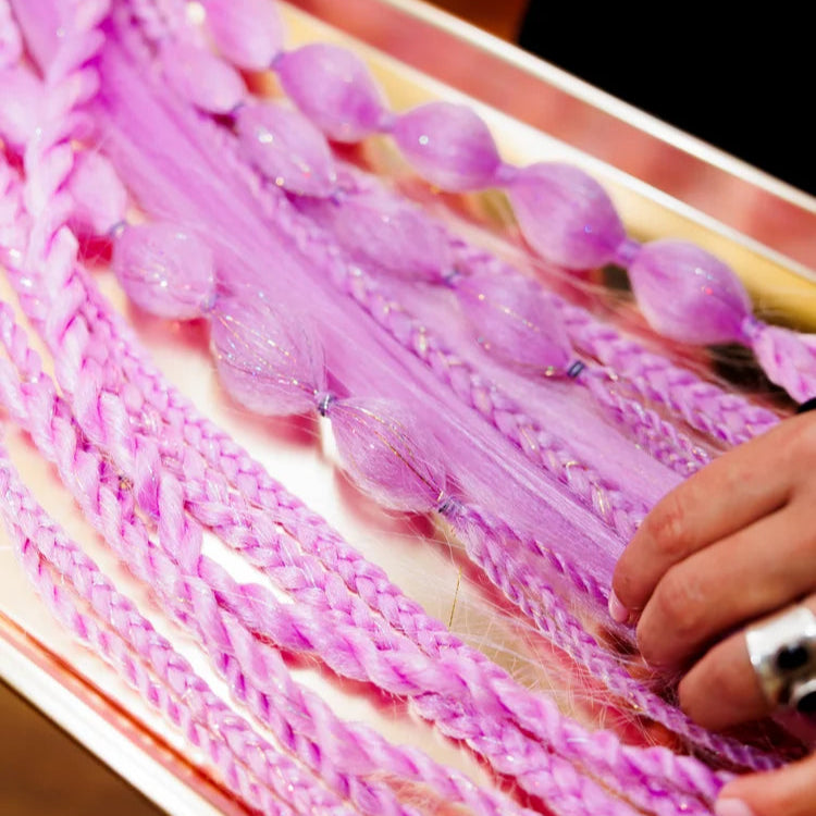 Close-up of a hand holding pink braided hair extensions on a wooden board.