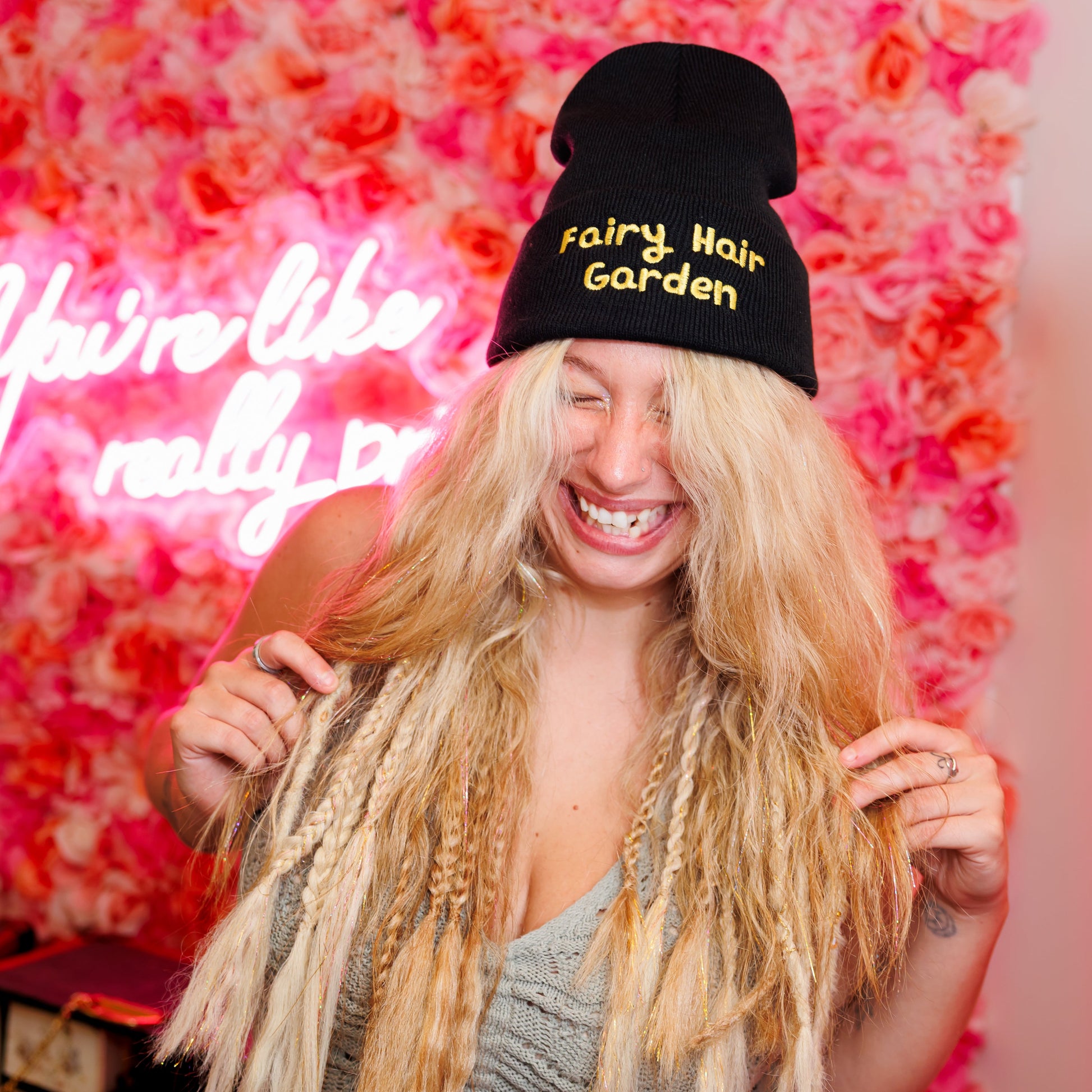 Person with blonde hair and a black beanie with 'Fairy Hair Garden' text in front of a pink floral wall and neon sign.