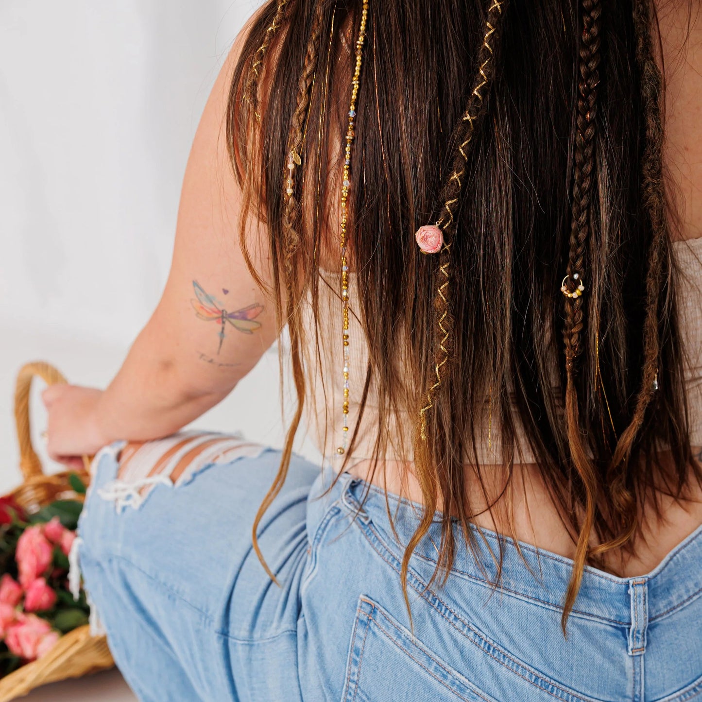 Back view of woman with boho braids, beads, and golden threads, holding rose-filled basket, dragonfly tattoo visible